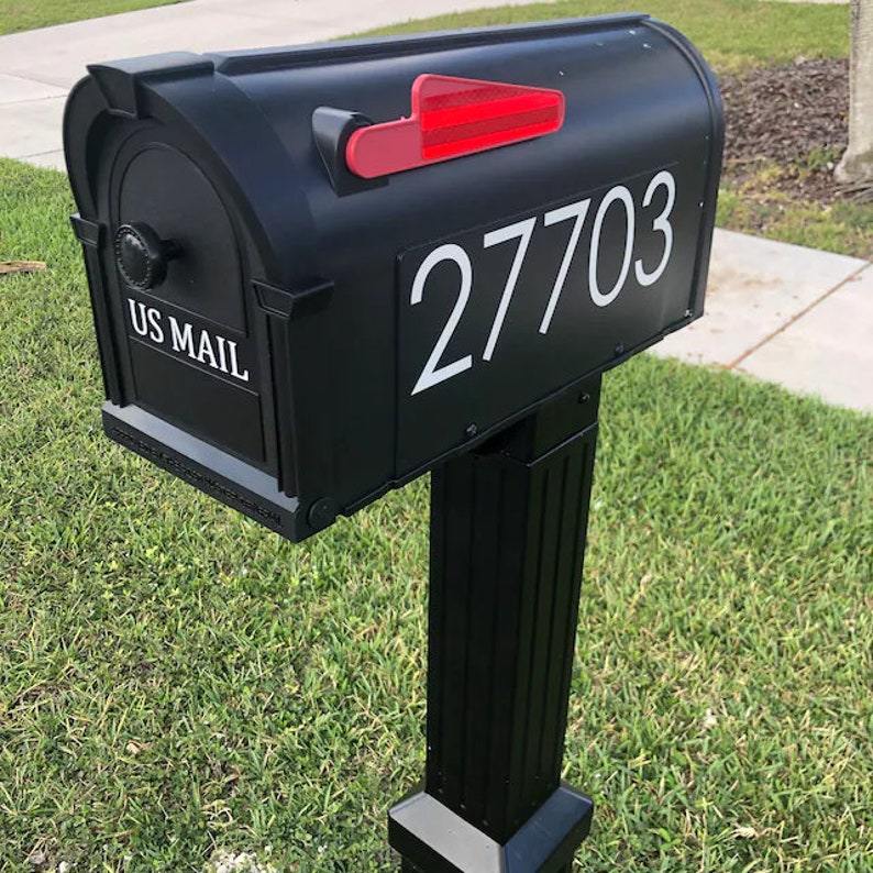 May include: Black mailbox with a red flag and the number 27703 on the front. The mailbox is mounted on a black post and is surrounded by green grass.