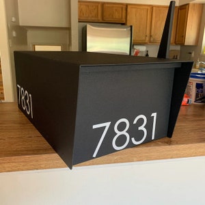 May include: Black mailbox with a white number "7831" on the front. The mailbox is sitting on a wooden surface.