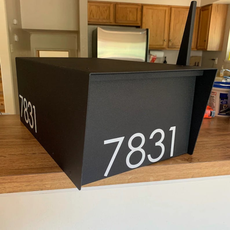 May include: Black mailbox with a white number "7831" on the front. The mailbox is sitting on a wooden surface.