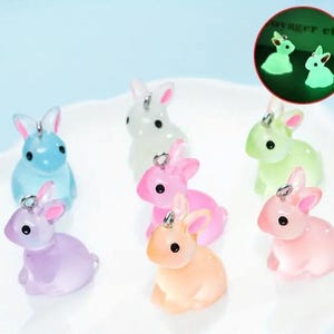 May include: A collection of colorful, translucent bunny charms in various pastel shades, including blue, pink, and lavender. Each bunny has a small silver loop for attaching to jewelry or other crafts. A glowing green bunny is also shown.