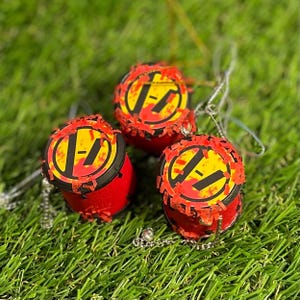 May include: Three red and yellow drum-shaped ornaments with a black and yellow logo, on green grass. Each ornament has a silver chain. The handmade ornaments have a unique, textured appearance. The design is eye-catching.