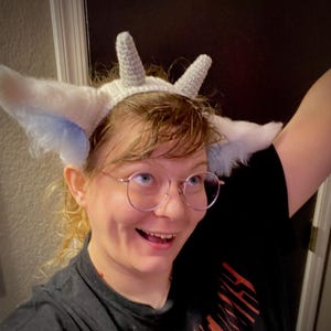 May include: A white crocheted headband featuring two pointed, textured horns on top and large, fluffy white ears with light blue accents on the sides. The headband is constructed with a dense stitch pattern and soft, faux fur material for the ears.