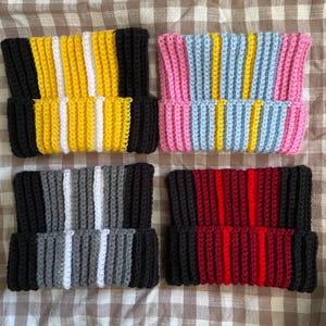 Peut inclure: Quatre bonnets crochetés faits à la main dans diverses combinaisons de couleurs. Un bonnet présente des rayures jaunes, blanches et noires. Un autre a des rayures roses, bleues et jaunes. Les deux autres bonnets ont des rayures grises et noires, et rouges et noires.