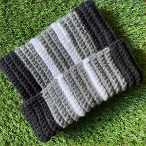 May include: A handmade crochet beanie in a striped pattern of black, white, and gray. The hat is folded over at the brim, and the texture of the yarn is visible. The beanie is displayed on a green surface.