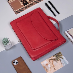 May include: A red leather portfolio with a zipper closure and a handle. The portfolio has a large pocket on the front and the word "DESIGN" is visible on a brown and black patterned notebook in the background.