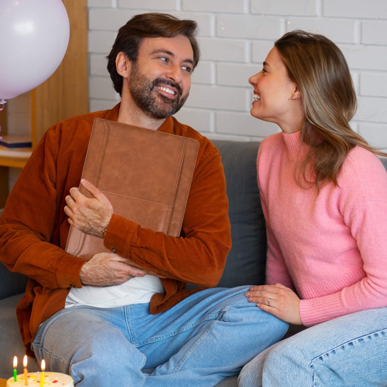 K&ouml;nnte beinhalten: Ein Mann und eine Frau sitzen auf einem Sofa. Der Mann h&auml;lt ein braunes, ledergebundenes Buch. Die Frau l&auml;chelt und schaut den Mann an. Im Vordergrund steht ein Geburtstagskuchen mit Kerzen.