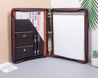 May include: A brown leather binder with a zipper closure, three pockets, and a pen holder. The binder is open and has a white sheet of paper inside. The binder has a silver ring mechanism.