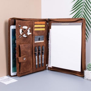 May include: Open brown leather portfolio with a zip closure. The interior includes a tablet, card slots, pen holders, and a notepad. A white charging cable is secured with leather straps. The portfolio is open, displaying its contents.