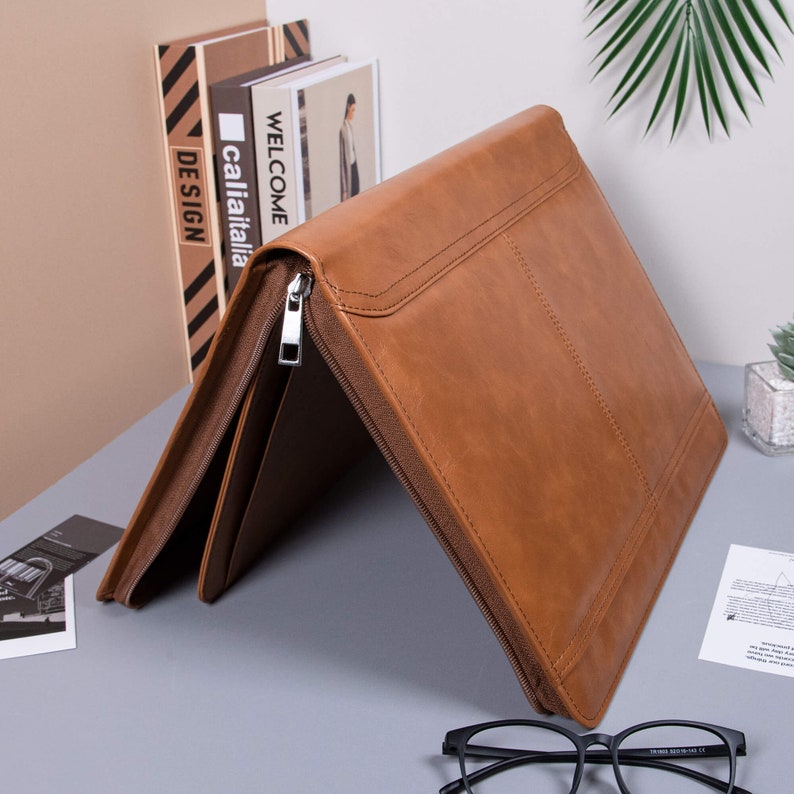 K&ouml;nnte beinhalten: Ein braunes Leder-Zipper-Portfolio-Ordner mit einem silbernen Rei&szlig;verschluss. Der Ordner ist geschlossen und liegt auf einer grauen Oberfl&auml;che.