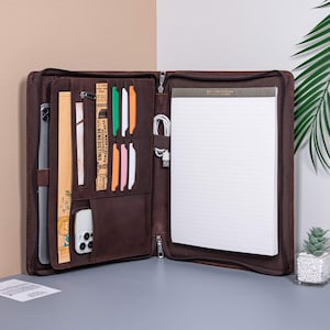 May include: A brown leather zippered portfolio with multiple pockets and slots for holding papers, pens, and a phone. The portfolio is open to reveal a lined notepad and a few colorful dividers.
