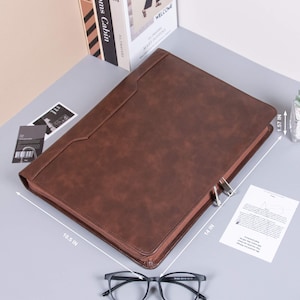 Op de afbeelding: Een bruine leren portfolio met ritssluiting, 35,56 cm lang, 26,67 cm breed en 3,99 cm dik. De portfolio is open en toont een paperclip en een document. Een bril ligt op de voorgrond.
