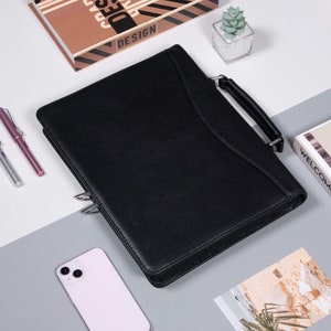 May include: A black leather portfolio with a silver zipper and a handle. The portfolio is lying on a gray surface with a white phone and a book with the word "DESIGN" on the cover.