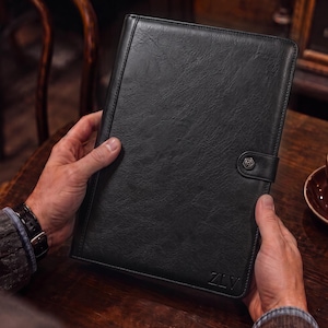 May include: A black rectangular portfolio with a textured surface, featuring a vertical stitched seam on the left side, a snap button closure on the right, and the letters ZLV embossed in the bottom right corner of the front cover.
