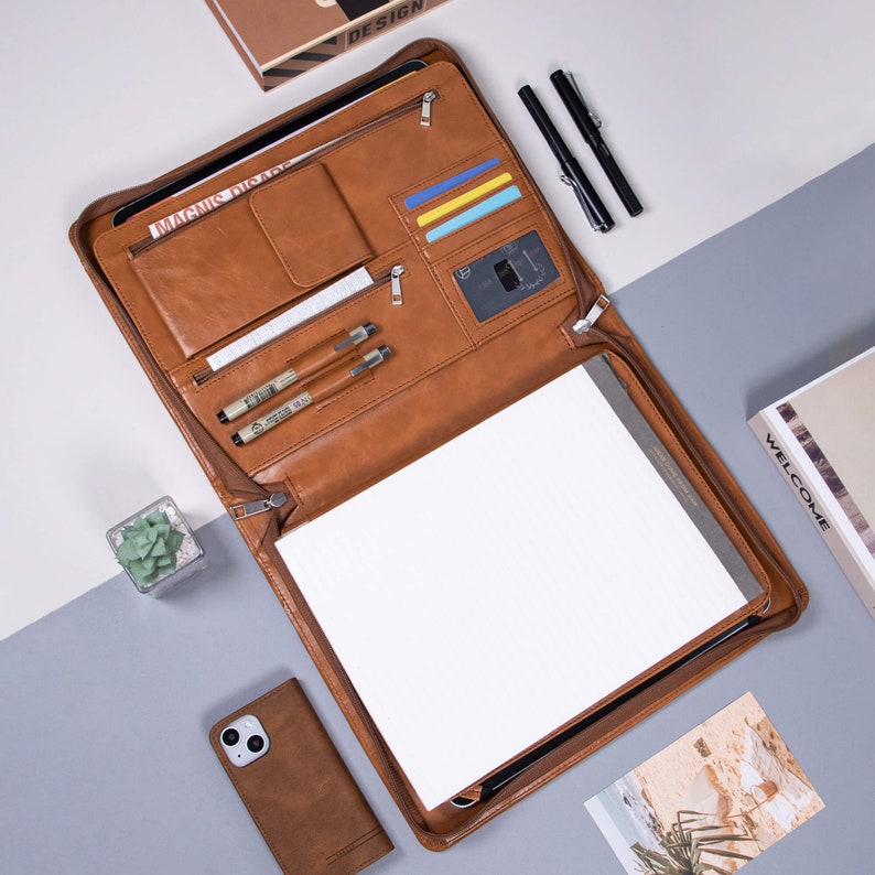 K&ouml;nnte beinhalten: Ein braunes Lederportfolio mit Rei&szlig;verschluss und mehreren Taschen. Das Portfolio enth&auml;lt einen Notizblock, Stifte und Kartenf&auml;cher. Der Text "MAGNIFIC" ist auf der Vorderseite des Portfolios sichtbar.