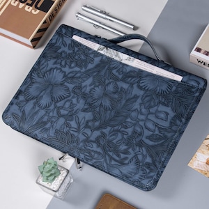 May include: Blue leather portfolio with embossed floral design. Features a handle, zipper closure, and a pocket. Includes a small succulent plant in a glass cube and two silver pens.