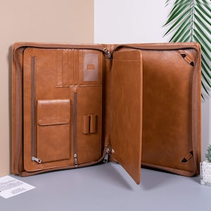 K&ouml;nnte beinhalten: Eine braune Ledermappe mit mehreren Taschen und einem Rei&szlig;verschluss. Die Mappe ist ge&ouml;ffnet und zeigt eine gro&szlig;e Tasche im Inneren.