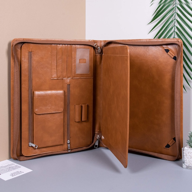 K&ouml;nnte beinhalten: Eine braune Ledermappe mit mehreren Taschen und einem Rei&szlig;verschluss. Die Mappe ist ge&ouml;ffnet und zeigt eine gro&szlig;e Tasche im Inneren.