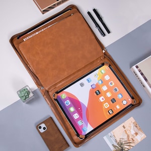 K&ouml;nnte beinhalten: Eine braune Ledermappe mit Rei&szlig;verschluss und einem integrierten St&auml;nder f&uuml;r ein iPad oder Tablet. Die Mappe ist ge&ouml;ffnet und das iPad ist sichtbar, wobei der Bildschirm verschiedene Apps anzeigt.