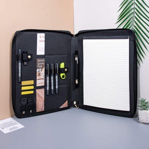 May include: Black leather portfolio with a zipper closure, a notepad, and a variety of pockets for pens, cards, and other accessories. The portfolio is open and the notepad is visible. The text "L'a sin" is visible on the inside of the portfolio.