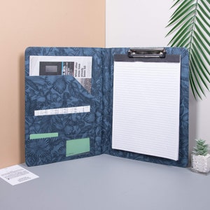 Könnte beinhalten: Ein blaues Clipboard mit floralem Muster, einem schwarzen Clip und einem weißen, linierten Notizblock. Das Clipboard hat mehrere Taschen zum Verstauen von Dokumenten und anderen Gegenständen.