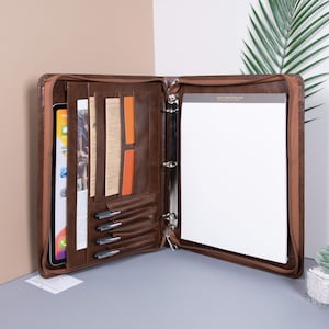 May include: Brown leather zippered binder with multiple pockets and a ring binder mechanism. The binder is open to reveal a white notepad and several pens.