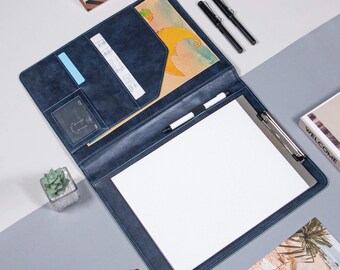 May include: A blue leather portfolio with a clipboard, a pen, and a small pocket on the front. The portfolio is open and has a white sheet of paper inside.