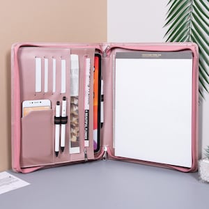 Pode incluir: Uma pasta de couro rosa com zíper com um bloco de notas, um tablet, canetas e um telefone dentro. O bloco de notas tem o texto "Ideas come from jam" na capa.