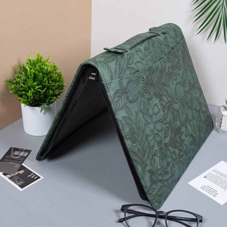 May include: A green leather portfolio with a floral embossed design. The portfolio has a zipper closure and a handle.