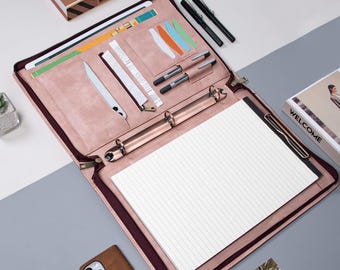 May include: A pink leather zippered binder with a gold ring mechanism. The binder has multiple pockets and slots for holding pens, cards, and other items. The binder is open to reveal a lined notepad.