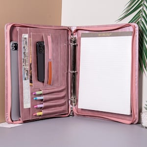 Pode incluir: Pasta rosa aberta de couro sintético com vários bolsos, um bloco de notas e uma pasta de três argolas. A pasta contém um tablet, um smartphone, canetas e outros itens. A pasta está aberta e pronta para uso.