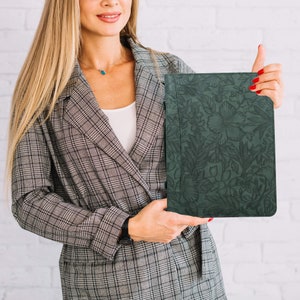 May include: A dark green leather folder with a floral embossed design. The folder is held by a person wearing a gray plaid blazer.