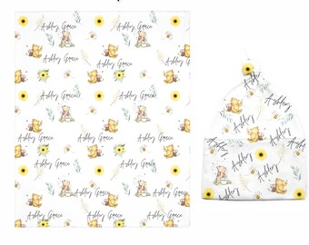 May include: A Winnie the Pooh themed baby gift set with a soft white fleece blanket featuring a repeating pattern of Winnie the Pooh, Piglet, bees, and sunflowers. The blanket is 30 inches by 40 inches. The set also includes a white knotted beanie with a similar pattern. The beanie and blanket are personalized with the name "Ashley Grace".