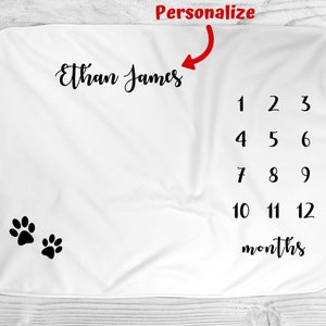 May include: White baby blanket with black paw prints and a personalized name, "Ethan James", and numbers 1-12 for monthly milestones.