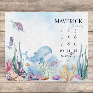 May include: A blue whale, seahorse, and turtle are featured on this baby milestone growth chart. The chart has a watercolor background with coral and seaweed. The numbers 1 through 12 are printed on the chart, along with the word "months". The name "Maverick James" is printed at the top of the chart.