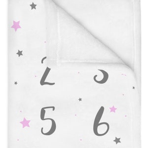 Puede incluir: Manta de beb&eacute; blanca con un patr&oacute;n de estrellas grises y rosas y n&uacute;meros grises del 1 al 9 impresos en ella.