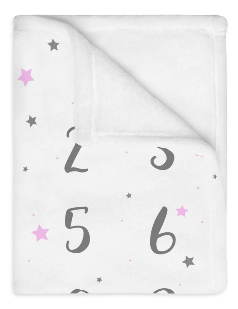 Puede incluir: Manta de beb&eacute; blanca con un patr&oacute;n de estrellas grises y rosas y n&uacute;meros grises del 1 al 9 impresos en ella.