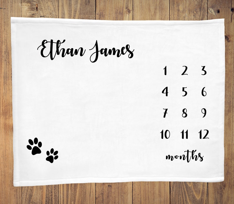 May include: White baby milestone blanket with black paw prints and the text "Ethan James" and numbers 1-12 with the word "months" below.