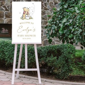 May include: White welcome sign with a Winnie the Pooh illustration, gold lettering, and the text "Welcome to Evelyn's Baby Shower 10.14.20XX".