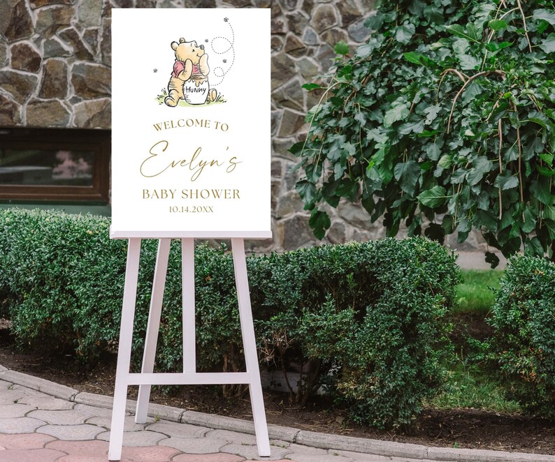 May include: White welcome sign with a Winnie the Pooh illustration, gold lettering, and the text "Welcome to Evelyn's Baby Shower 10.14.20XX".