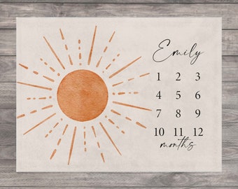Puede incluir: Un gráfico de crecimiento para bebé beige con un gráfico de sol en naranja y marrón. El gráfico tiene los números del 1 al 12 para cada mes del año. El nombre "Emily" está escrito en la parte superior del gráfico.