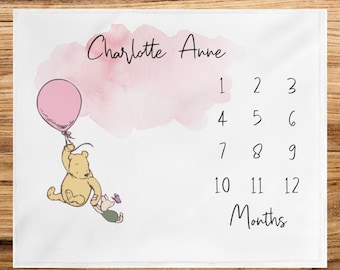May include: A white baby milestone blanket with a pink watercolour background. Winnie the Pooh and Piglet are holding hands and floating in the air with a pink balloon. The text "Charlotte Anne" is at the top of the blanket. The numbers 1 through 12 are listed in three rows, with the word "Months" below the numbers.