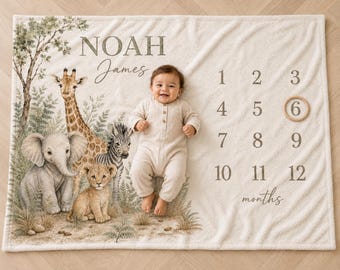 May include: A cream-colored milestone blanket featuring a watercolor illustration of a giraffe, elephant, zebra, and lion cub. The text reads NOAH James and months 1 through 12. A wooden ring circles the number 6 on the soft, textured fabric.