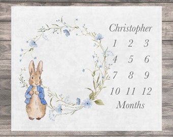 May include: A white milestone blanket featuring a watercolor illustration of a brown rabbit wearing a blue jacket next to a floral wreath. The text reads Christopher, the numbers 1 through 12, and the word Months in a grey serif font.