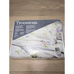 May include: A Trousseau brand bedding set package featuring white fabric with a floral pattern of purple and blue flowers with green stems. The plastic packaging includes printed text, care instructions, and a barcode label on the front.