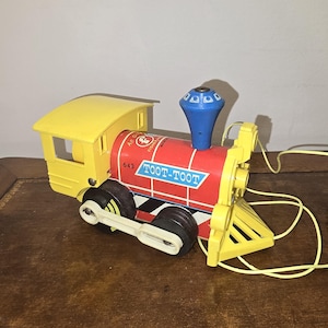 May include: A vintage toy train engine featuring a yellow cab and cowcatcher, a red cylindrical boiler with the text TOOT-TOOT and 643, a blue smokestack, and black wheels with white connecting rods. A yellow pull string is attached to the front.