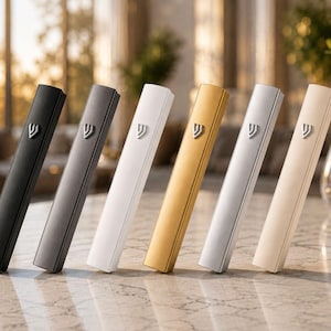 May include: A set of six rectangular mezuzah cases standing upright in a row. The cases are finished in matte black, dark gray, white, gold, silver, and cream. Each case features a silver-colored Hebrew letter Shin centered on the upper portion.
