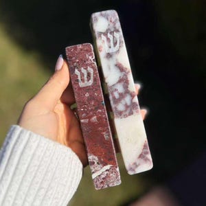 Puede incluir: Dos estuches rectangulares de piedra para mezuzá. Uno es rojo intenso con vetas blancas, el otro es blanco con vetas rojas. Cada uno tiene una letra hebrea plateada en la parte delantera. Sobre un fondo borroso verde y marrón.