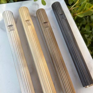 Puede incluir: Cuatro estuches de mezuzá metálicos en plata, oro, bronce y negro. Cada estuche tiene un diseño acanalado y una pequeña letra grabada. Los estuches son cilíndricos y descansan sobre una superficie de mármol blanco.