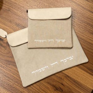 May include: Two beige fabric pouches with smooth, light-colored flaps. Each pouch features white embroidered Hebrew text that reads "יְבָרֶכְךָ ה' וְיִשְׁמְרֶךָ". The pouches are rectangular with a textured weave, and one is significantly larger than the other.
