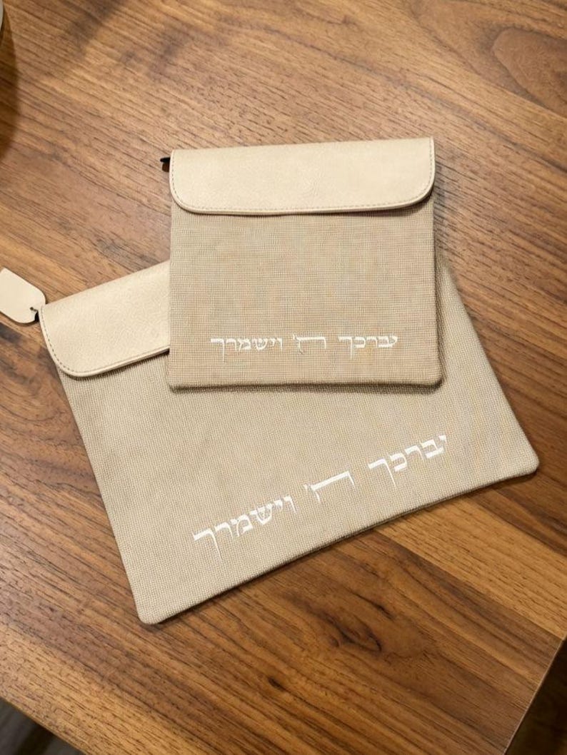 May include: Two beige fabric pouches with smooth, light-colored flaps. Each pouch features white embroidered Hebrew text that reads "יְבָרֶכְךָ ה' וְיִשְׁמְרֶךָ". The pouches are rectangular with a textured weave, and one is significantly larger than the other.